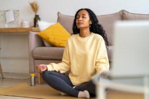 Frau in Sportkleidung mit Bärenfüßen sitzt im Lotussitz auf einer Matte im Wohnzimmer, während sie meditiert und Atemübungen macht. Konzept der Achtsamkeit und des Stressabbaus.