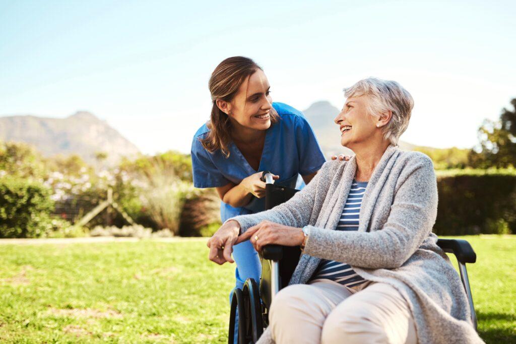 Eine Pflegekraft in blauer Uniform lächelt eine ältere Frau im Rollstuhl an. Die beiden befinden sich draußen in einem sonnigen Garten mit Bergen im Hintergrund.