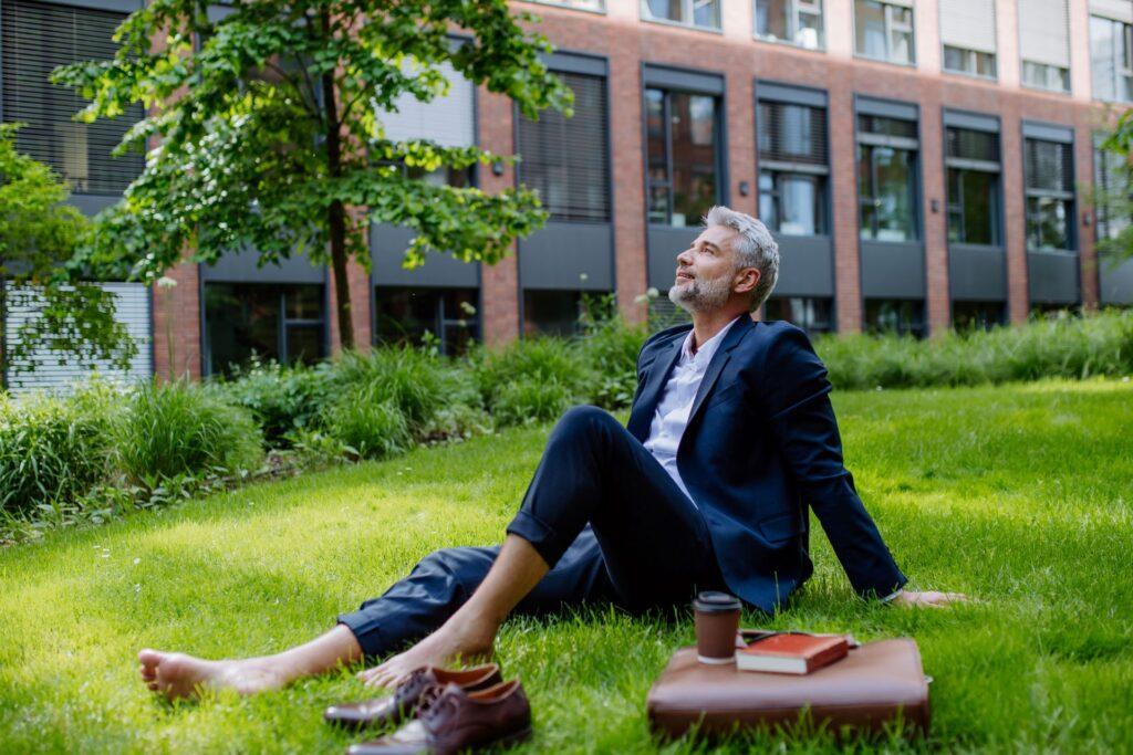 Barfüßiger Geschäftsmann im Anzug sitzt entspannt auf einer Wiese, lehnt sich zurück und genießt seine Pause im Grünen vor einem modernen Bürogebäude.