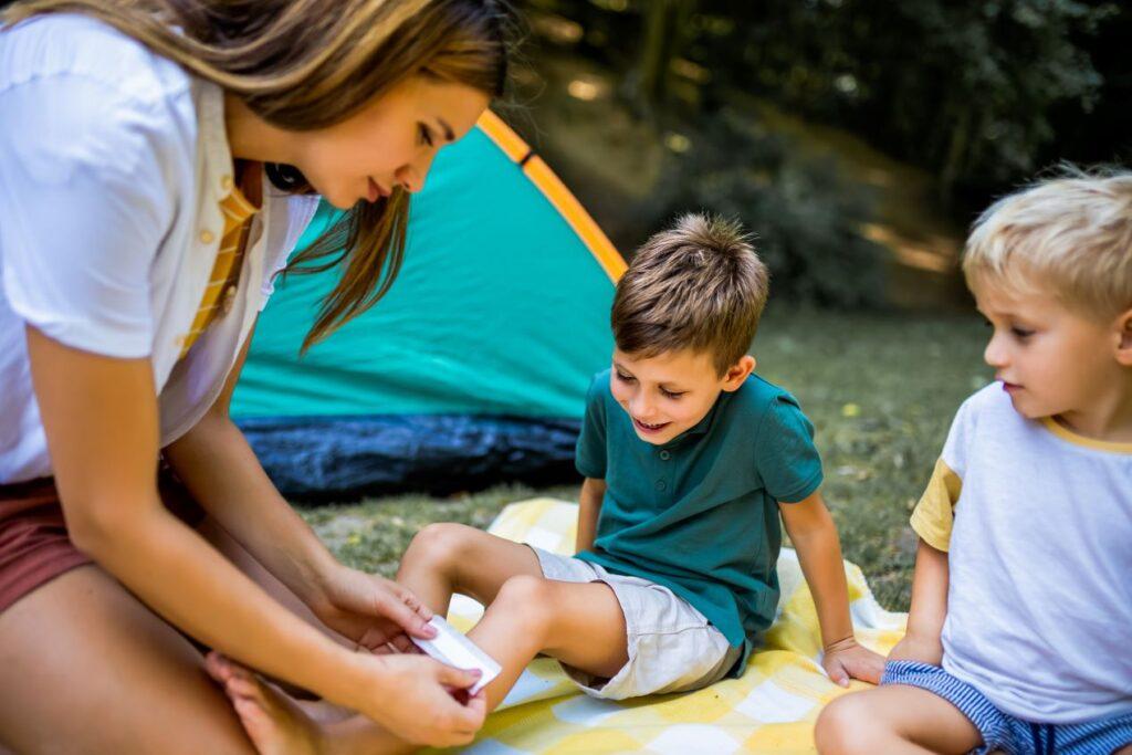 Eine Frau leistet Erste Hilfe und versorgt das Knie eines Kindes mit einem Verband während eines Campingausflugs im Freien, neben einem zweiten Kind und einem Zelt.