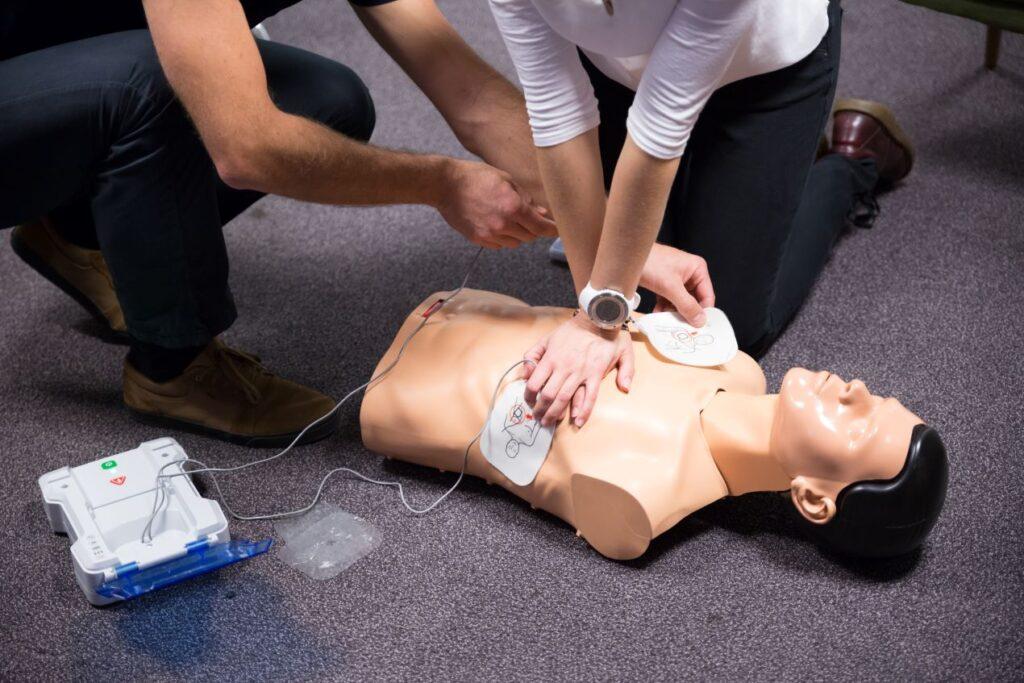 Teilnehmer der Nothelfer Instruktor Ausbildung üben die Anwendung eines Defibrillators und die Herz-Lungen-Wiederbelebung an einer Übungspuppe, um lebensrettende Maßnahmen zu trainieren.