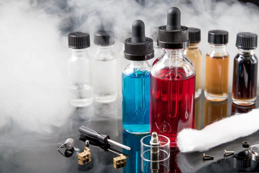 Mehrere Flaschen mit bunten E-Liquids und Zubehör wie Coils, Watte und Schrauben für E-Zigaretten auf einer spiegelnden Oberfläche mit Dampf im Hintergrund.