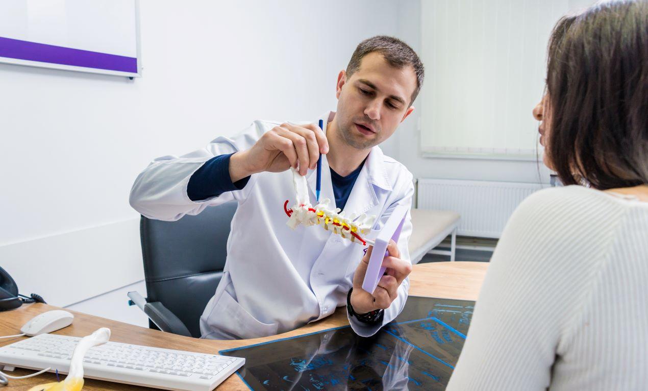 Ein Orthopäde erklärt einer Patientin mit einem Modell der Wirbelsäule, wie ein ergonomisches Boxspringbett Rückenschmerzen lindern kann.