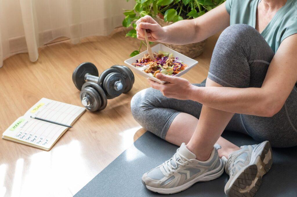 Frau sitzt auf Yogamatte mit gesundem Essen, Kurzhanteln und 90-Tage-Trainingsplan